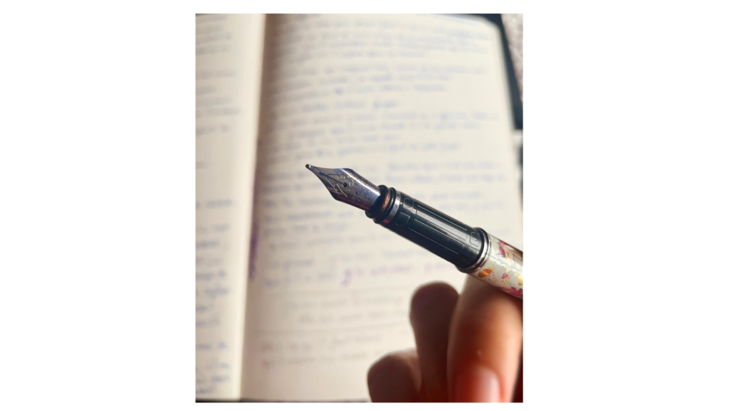 un stylo plume en premier plan tenu dans une main, en arrière plan un carnet